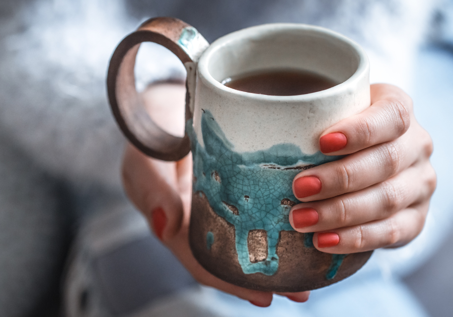 A ladies hands with burnt orange nail varnish holding a full cup of tea. The cup is rustic, with blue and bronze colouring.