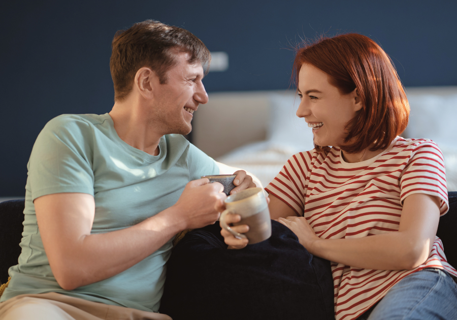 A man and women sitting on a sofa holding cups of tea smiling and talking to one another.