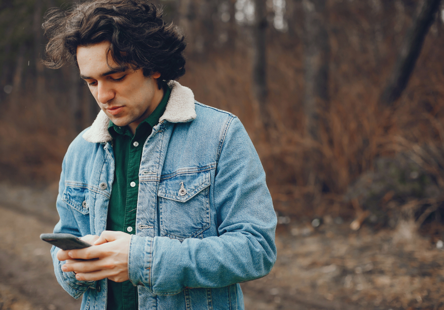 A man in a woody area looking at his mobile phone in his hand.